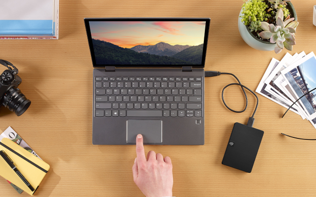 Overhead view of a person uses a laptop connected to a Seagate portable external drive, with a camera, glasses, and printed photos nearby.