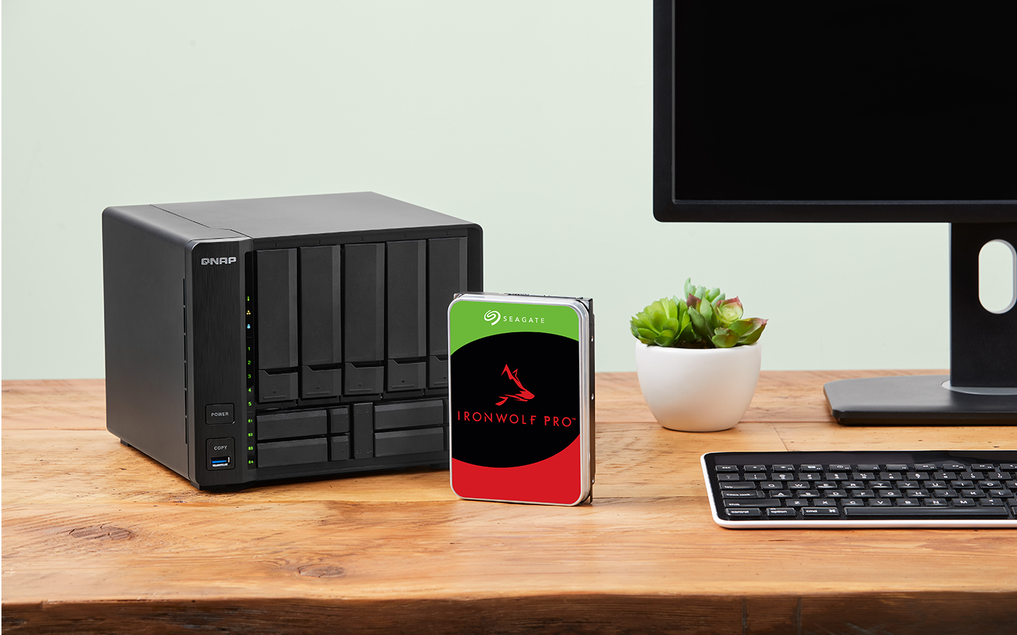 IronWolf Pro drive positioned centrally on a desk is surrounded by a NAS system, computer screen, and keyboard.