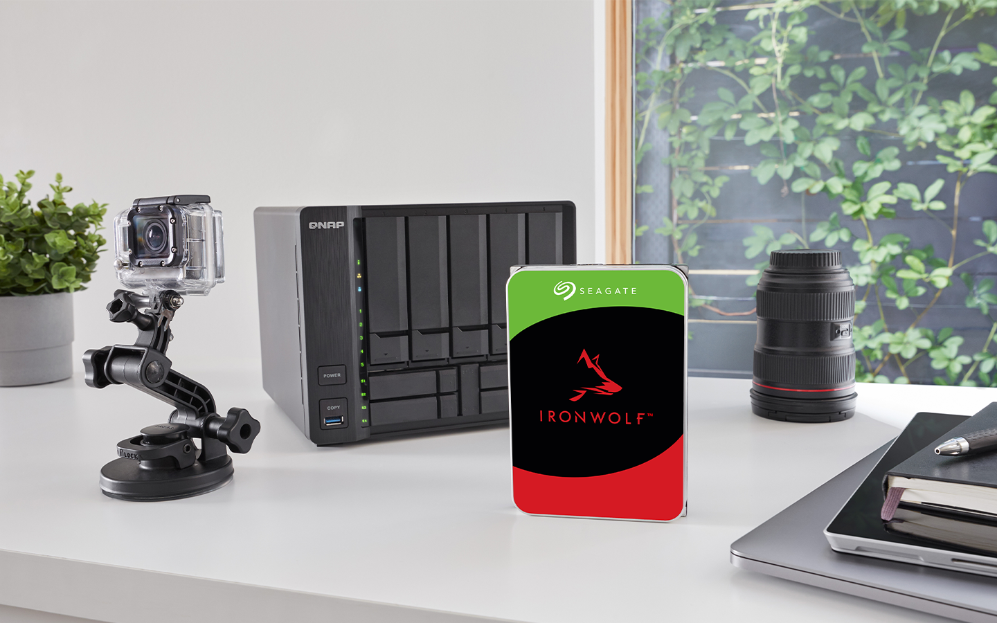 A FireCuda NAS drive stands before a digital camera setup, NAS system, camera lens, and stacked laptop, tablet, and notebook.