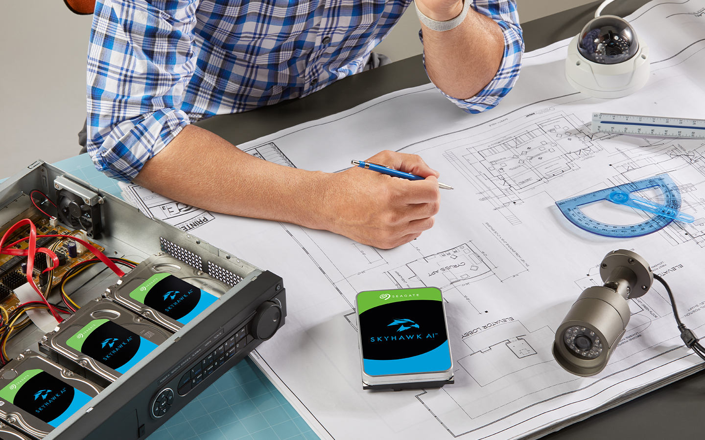 A person sits at a desk looking at a building blueprint with two surveillance cameras and four Seagate Skyhawk AI storage drives.