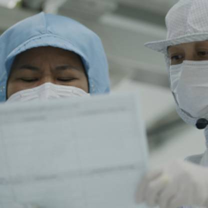 Two tech lab workers wearing full-body clean room gear review information on paper they are holding.