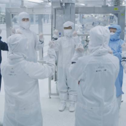 A group of nine people wearing clean room, full-body suits stand in a circle in tech lab.