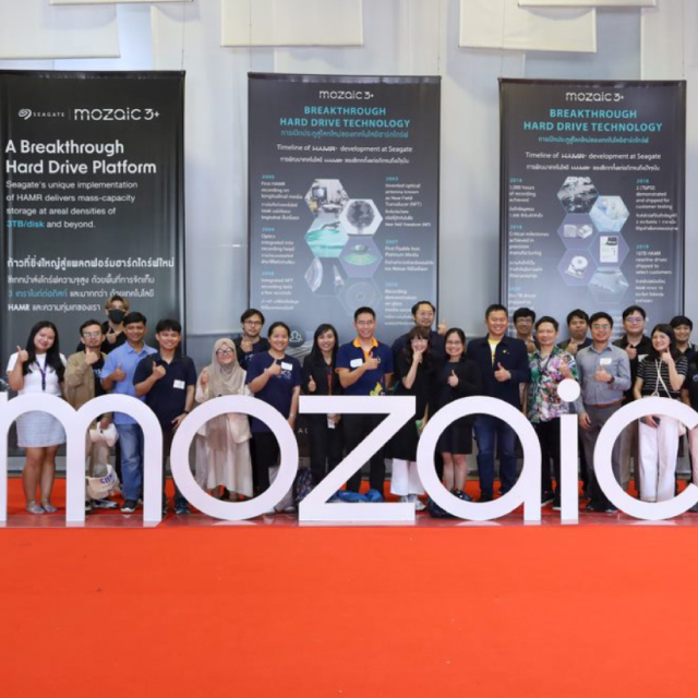Group shot of smiling people giving a thumbs up and standing behind a large dimensional white Mosaic3+ logo with a series of oversized Mosaic3+ banners behind them.