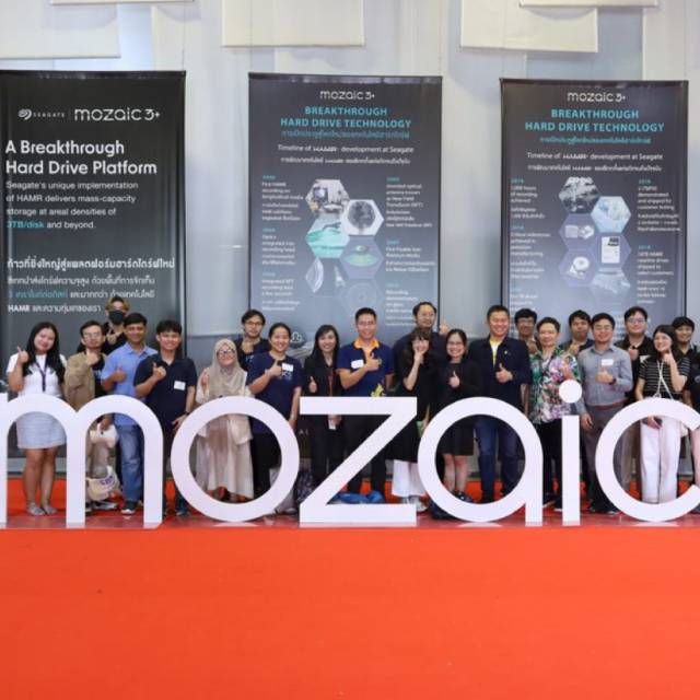 Group shot of smiling people giving a thumbs up and standing behind a large dimensional white Mosaic3+ logo with a series of oversized Mosaic3+ banners behind them.