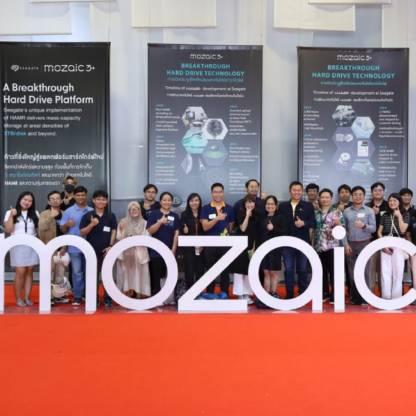 Group shot of smiling people giving a thumbs up and standing behind a large dimensional white Mosaic3+ logo with a series of oversized Mosaic3+ banners behind them.