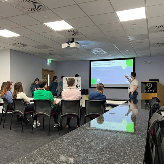 An employee presenter in a Seagate conference room stands to the right of a projection screen and points to a slide for a small group of students.