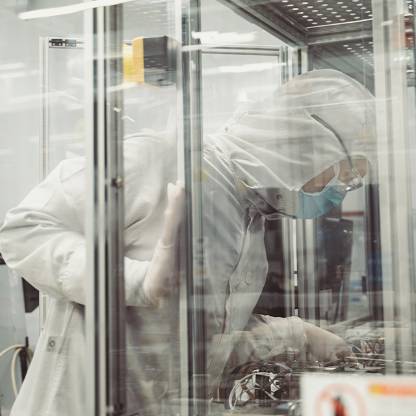 A person dressed in a clean room suit leans into a glass case to review technology.