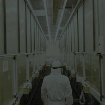 A person wearing a full-body clean room suit stands in an aisle with shelves of shrink-wrapped cardboard boxes on both sides.