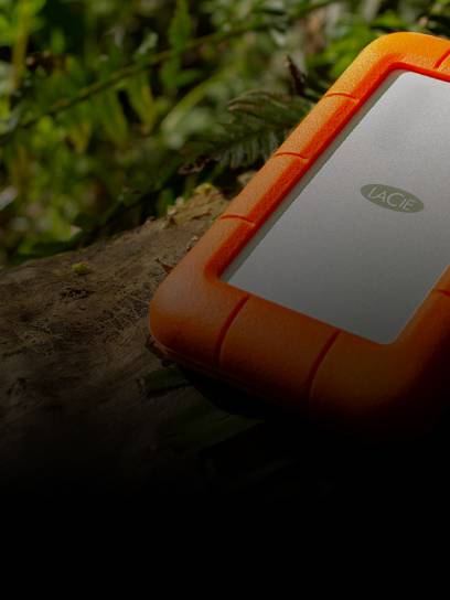 Featuring an orange protective cover, a LaCie Rugged Mini drive sits on a log in the outdoors next to a backpack.