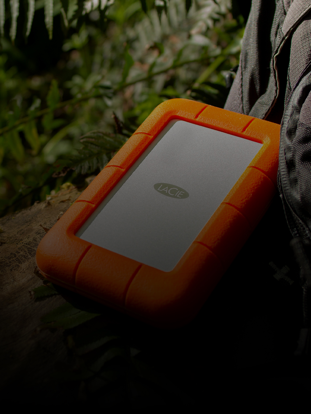 LaCie Rugged USB-C external hard drive sits next to a backpack on rocks, showing its durability when used outdoors.