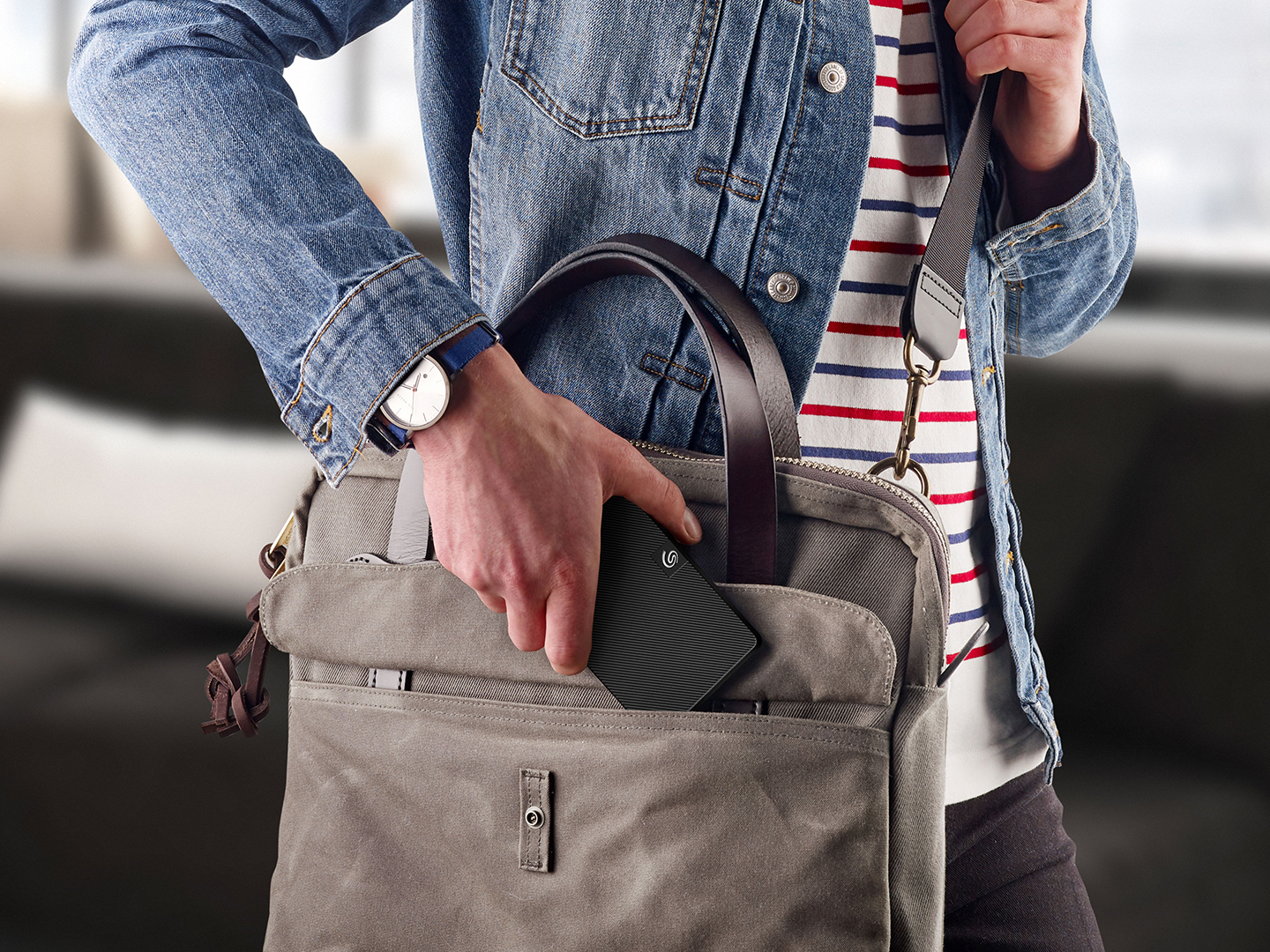 <add alt text>A person putting a Seagate portable external drive into the open pocket of a gray messenger bag being carried.