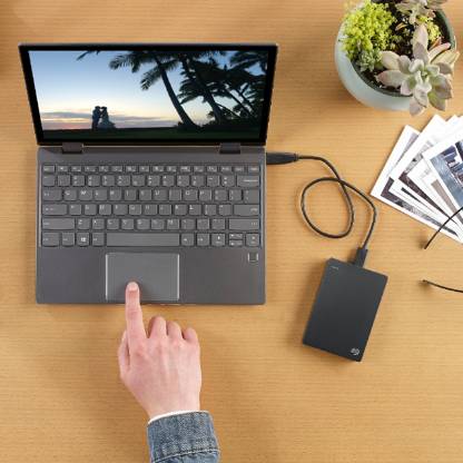 A small, black Seagate external hard drive connected to a laptop that features a photo of a couple next to trees. The user has a finger on the laptop’s touchpad and a pair of glasses, additional printed images, and a small planter of succulents are featured next to the laptop.