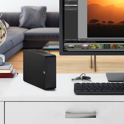 A home office setup with a black Seagate desktop drive next to a keyboard and flat screen display. Near the drive, a set of keys and a few books are displayed, and on the screen is a series of photos.