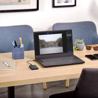 A portable external hard drive connected to a laptop sits on a wooden desk with a notepad, pen cell phone, plant and sunglasses.